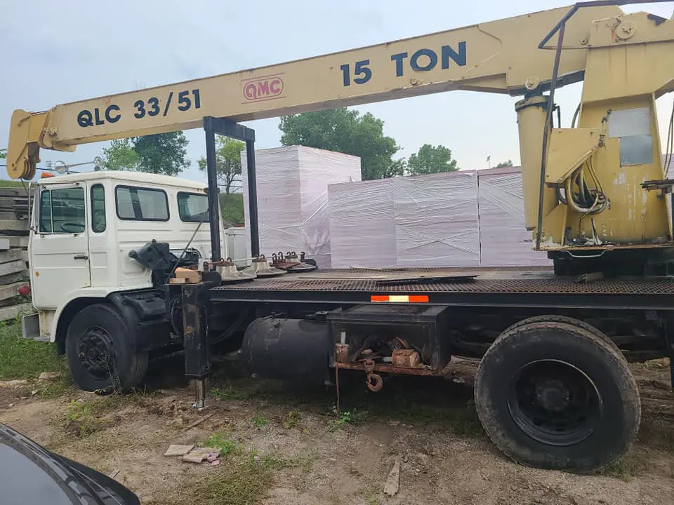 White flatbed service truck with a yellow 15 TON crane arm mounted on the bed, parked on dirt in front of wrapped pallet stacks, showing cab, outriggers and worn tires — illustrates onsite diesel repair and mobile equipment service.