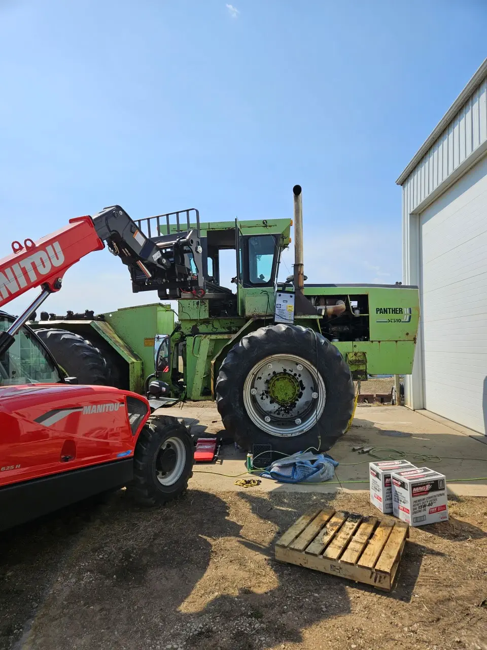 Large green Panther agricultural machine with a removed wheel being serviced onsite beside a white shop door, a red Manitou telehandler positioned to assist, cardboard boxes of parts and tools on the ground, clear blue sky — depicting onsite roadside diesel and equipment repair.