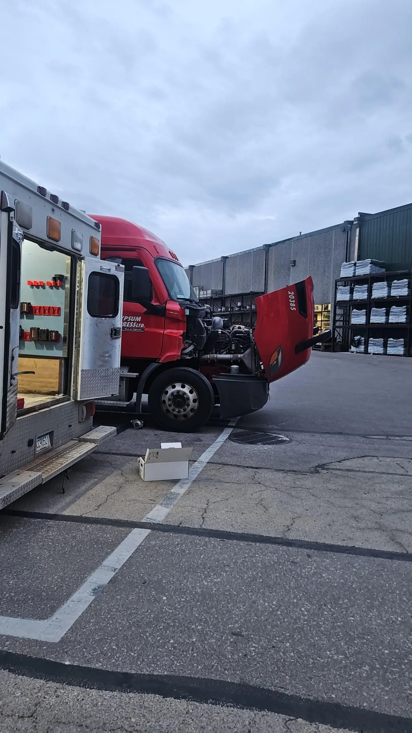 red semi truck cab with raised hood parked beside an open service truck at a warehouse loading area under overcast sky, mechanic tools visible in service truck and stacked pallets in background